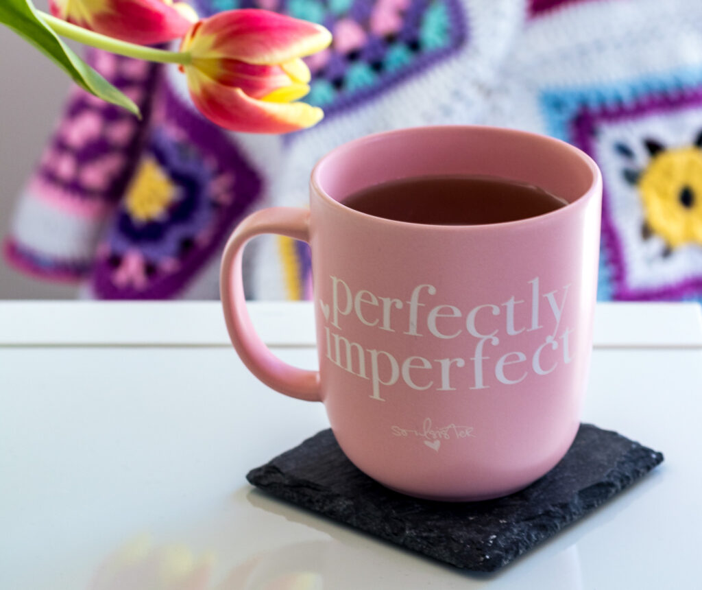 Teetasse mit der Aufschrift perfectly imperfect. Im Hintergrund Tulpen und eine bunte Häkeldecke